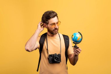 Düşünceli orta yaşlı erkek gezgin küçük bir dünya tutuyor, nereye gideceğini düşünüyor ve bakıyor, sarı arka planda sırt çantası ve fotoğraf makinesiyle duruyor.