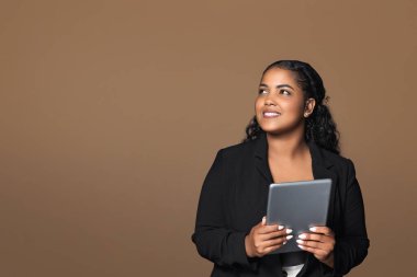 İş ilanı. Neşeli Brezilyalı vücut pozitifliği. Dijital tablet kullanan, kahverengi arka plan üzerinde duran, boş alana bakan iş kadını. Girişimci kadın aygıtla web sörfü yapıyor