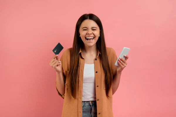 流动购物应用 快乐少女持信用卡和智能手机 兴奋快乐少女站在粉红工作室背景上 快乐少女在线购物的肖像 — 图库照片