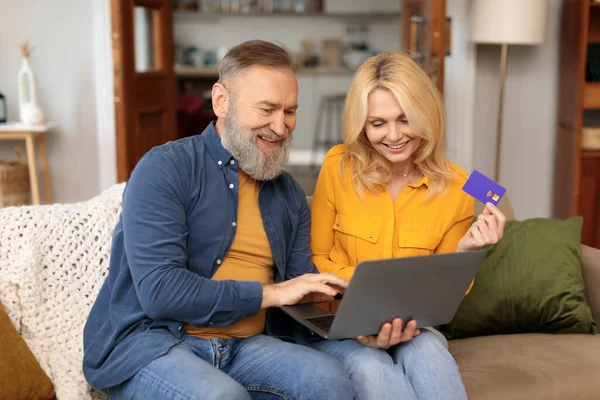 Internet Shopping, Ecommerce. Older couple buying something online, using laptop and credit card, sitting on couch at home. Man points at computer screen while wife holds bank card, making payment