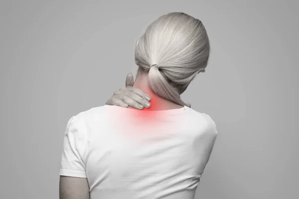 Rear view of tired mature woman touching inflamed zone on her neck, suffering from cervical osteochondrosis, massaging and rubbing sore area lightened with red, monochrome shot, copy space