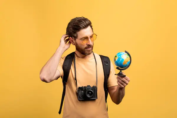 Stock image Thoughtful middle aged male traveler holding small globe, looking and thinking where to go next, standing with backpack and photocamera on yellow studio background