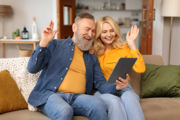 Seniors Virtual Communication. Happy Mature Couple Makes Video Call Using Digital Tablet Computer, Gesturing Hello Greeting And Communicating Online Sitting Together On Couch At Home