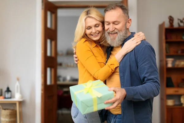 Happy holidays concept. Thankful senior caucasian woman hugging husband with gift box at cozy home. Mature couple celebrating birthday or anniversary holiday together. Valentines day present