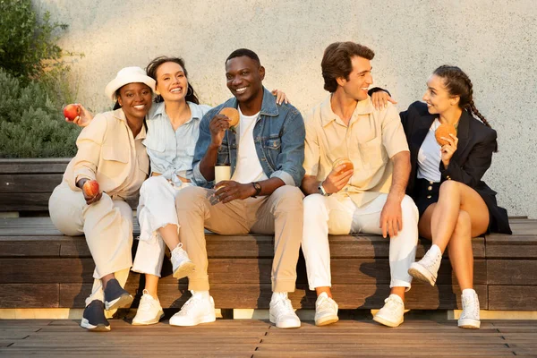 Multicultural group of friends cheerful young men and women sitting on bench at universtiry campus or at park, have lunch break outdoor, eating sandwiches, fruits, drinking takeaway coffee, chatting