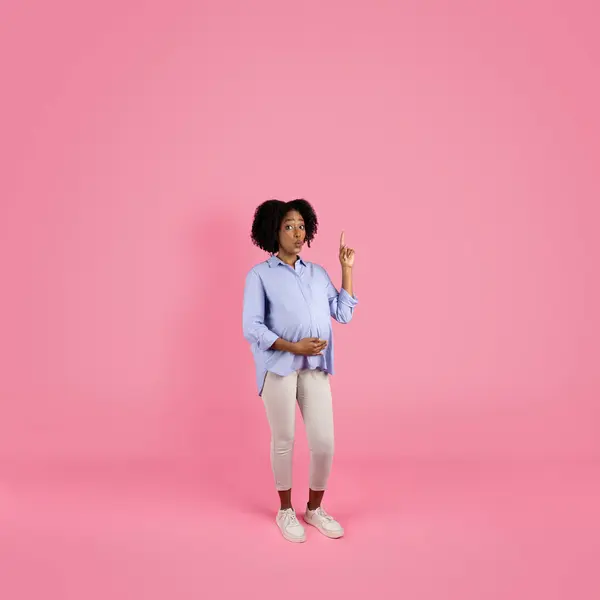 Smiling shocked inspired young black pregnant woman in casual touching big belly with hand, pointing finger at empty space, isolated on pink studio background. Got idea, ad and offer, waiting baby