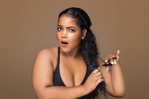Shocked brazilian chubby lady cutting her curly long hair, sad young woman unwilling to do haircut, posing on brown studio background, copy space. Hairdressing and unruly hair concept