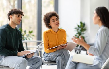 Üç uluslararası grup arkadaşı projelerini tartışıyor, üniversite sınıfında daireler çizerek oturuyorlar, araştırma ve fikir alışverişi yapıyorlar.