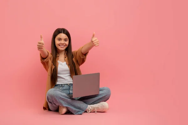 カメラで親指を示すラップトップと幸せなティーンガールの肖像画 新しいウェブサイトやオンラインコースをお勧めの陽気な女性学生 ピンクのスタジオの背景に座って コピースペース — ストック写真