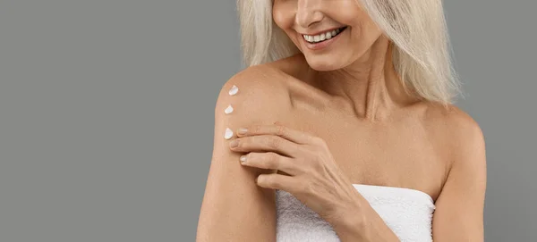 Anti-Aging Skincare. Smiling Mature Woman Applying Body Lotion On Shoulder, Unrecognizable Older Lady Moisturising Skin, Using Nourishing Cream While Standing Isolated On Grey Background, Copy Space