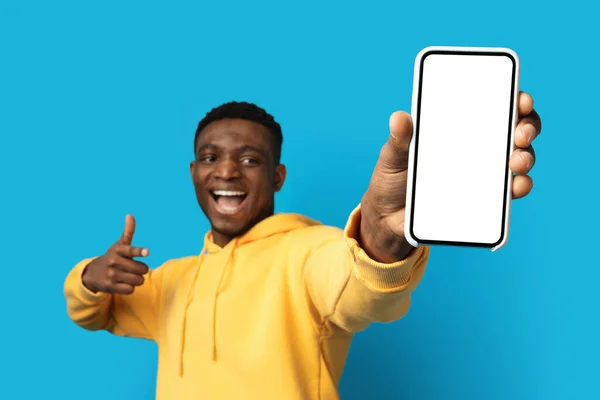 穿着黄色帽衫的冷静 情绪化的年轻的非洲裔美国人展示了一个大大的智能手机 屏幕为白色 大拇指翘起 背景为蓝色 推荐了一款漂亮的移动应用程序 — 图库照片
