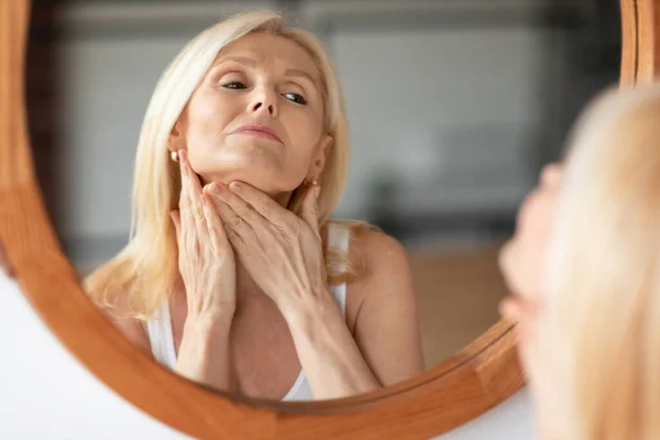 Double chin treatment. Middle aged european woman touching soft smooth skin on neck, looking at mirror in bedroom at home, enjoying domestic beauty routine