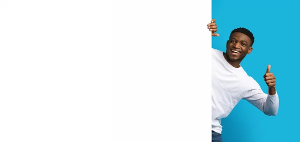 ポジティブな幸せなハンサムな若い黒人の男は横の白い広告板の隣にポーズし 親指を上げて微笑み 青いバックグランド モックアップ 空白のスペース バナーを表示します 素晴らしいオファー — ストック写真