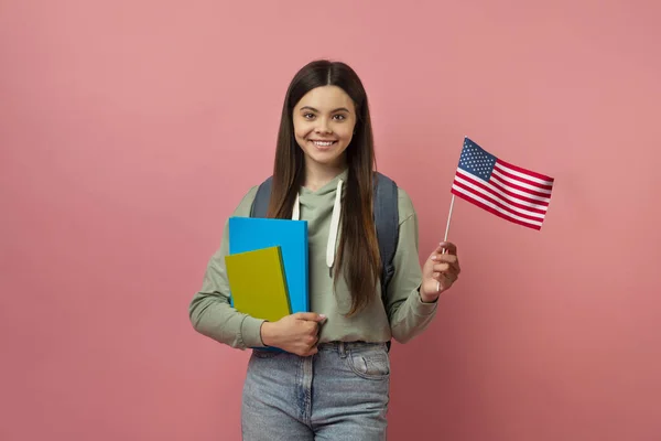 Happy teen exchange student girl with notebooks holding usa american flag, cheerful female teenager study english abroad, looking and smiling at camera, posing on pink studio background, copy space