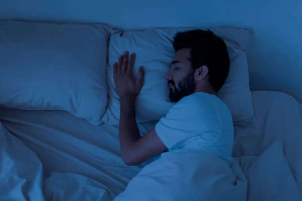 Peaceful handsome young indian man sleeping in comfortable bed alone at home, calm eastern guy enjoying his orthopedic mattress and pillow, resting in bedroom at night, having good sleep