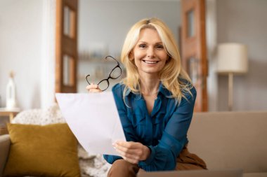 Evrak işleri. Elinde kağıtlar ve bardaklarla poz veren neşeli iş kadını portresi, evde çalışıyor, kameraya gülümsüyor. Mesafe çalışması, profesyonel danışma kavramı