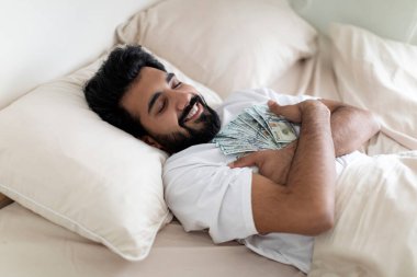 Mutlu Hintli genç adam elinde dolarlarla yatakta uyuyor, sakin, mutlu, elinde para banknotları tutan, gülümseyen, ekonomiden keyif alan, büyük kar getiren, boş alanla yakın çekim yapan...