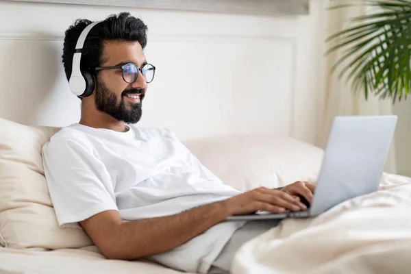 Smiling Indian Man In Wireless Headphones Typing On Laptop While Sitting In Bed, Happy Young Eastern Guy Working Remotely From Home, Using Computer For Online Communication, Side View, Copy Space