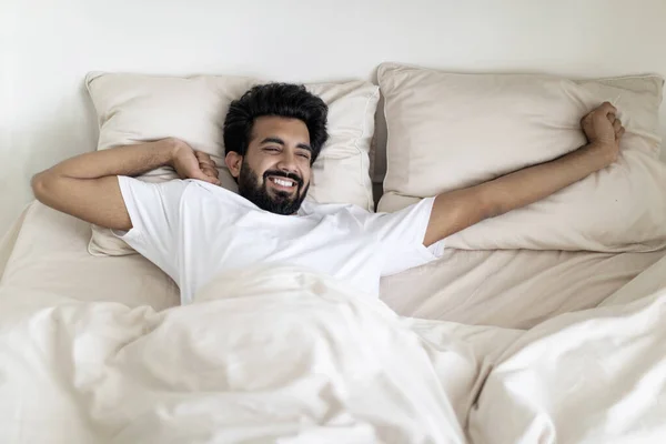 Lazy Morning. Happy Young Indian Man Stretching In Bed After Nice Sleep, Cheerful Millennial Eastern Guy Waking Up With Good Mood, Relaxing In Light Bedroom, Enjoying Weekend Pastime, Above View