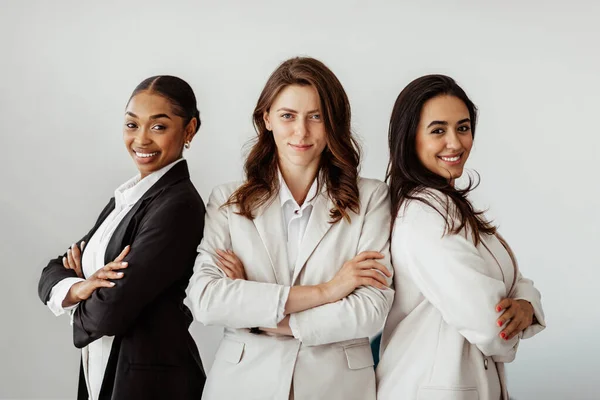 Three successful multicultural businesswomen standing back to back with arms crossed and smiling at camera, posing in office. Successful diverse female colleagues