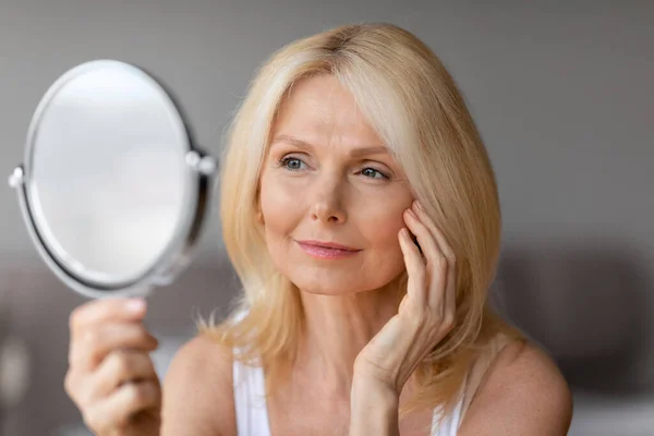 Beautiful middle aged blonde woman touching face and looking in mirror, enjoying her flawless skin, smiling female making anti-aging beauty routine, closeup