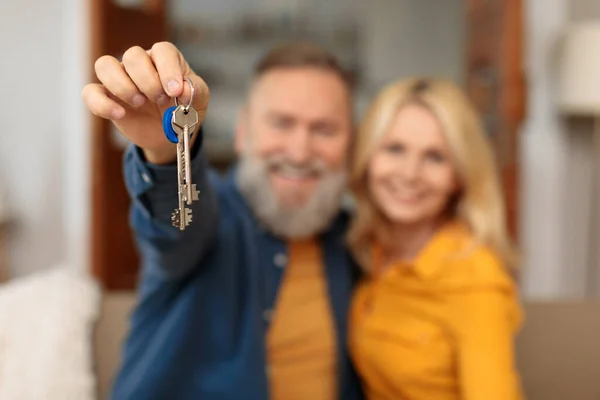 New house. Cheerful european senior couple showing keys in living room together, shallow depth. Mature husband and wife apartment owners advertising real estate mortgage or property rental offer