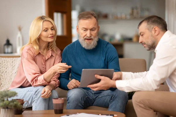 Real estate agent man showing house plans on electronic digital tablet to senior couple clients, looking at apartment floor plan, sitting on sofa indoor. Agency Service Offer