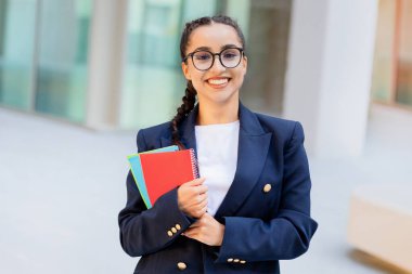 Neşeli, güzel, esmer, şık giyinmiş, gözlüklü bir bayan. Üniversite kampüsünde poz veren gözlüklü bir öğrenci. Elinde not defterleri var. Eğitim, okul, üniversite konsepti