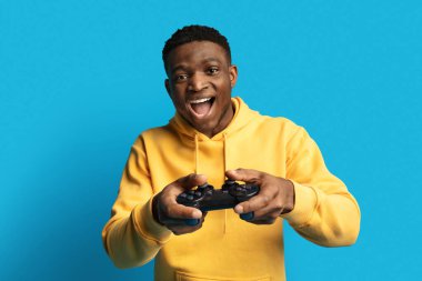 Mutlu, duygusal, Afro-Amerikalı bir adam, günlük kıyafetleriyle, mavi arka planda joystick ile video oyunları oynuyor, siyahi bir adam, kameraya bakıyor ve haykırıyor. Eğlence