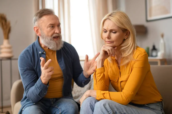 Couple Quarrel. Emotional mature man gesturing and shouting at his unhappy wife indoor, spouses having conflict sitting on couch at home. Relationship crisis, domestic abuse concept