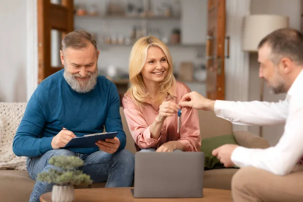 Real Estate Offer. Agent Man Giving Key To Senior House Buyers Couple Indoor, Cheerful Mature Spouses Buying Apartment Flat Sitting On Sofa And Signing Papers. Mortgage, New Property Purchase