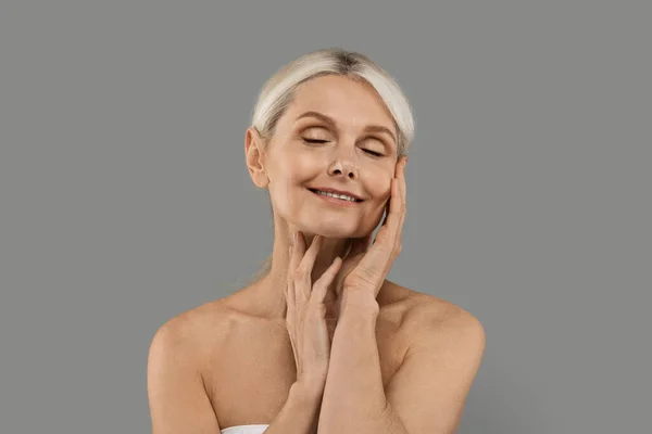 Anti-Aging Cosmetics. Senior Woman With Beautiful Soft Skin Posing Over Grey Background, Attractive Mature Female Touching Her Skin And Smiling, Enjoying Skincare Treatments, Copy Space