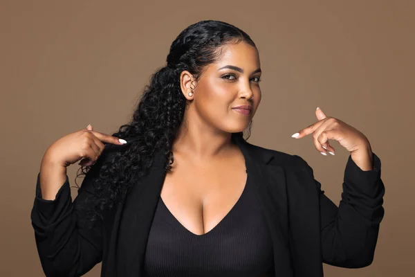 This is me. Self assured proud brazilian plus size woman pointing fingers indicating herself posing standing over brown background. Self-confidence concept