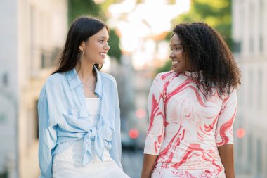 İki gülümseyen genç bayan arkadaş dışarıda birlikte rahatlıyor, mutlu çoklu etnik dostların portresi şehir sokağında sohbet ediyor ve dinleniyor, birbirlerine bakıyor, arkadaşlıklarının tadını çıkarıyorlar.