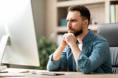 Ofiste çalışırken bilgisayar ekranına bakan odaklanmış genç iş adamının portresi, iş yerinde oturan dalgın milenyum girişimcisi, iş fikirleri üzerine kafa yoran,