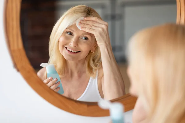 Happy middle aged woman looking at mirror and using micellar water with cotton pads, cleaning makeup and cosmetics after working day before shower