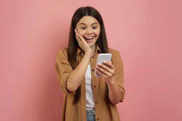 好消息 快乐兴奋的少女在智能手机上读信息 触动脸庞 用手机进行在线交流 站在粉色背景上 复制空间 惊讶地微笑着的少女 — 图库照片