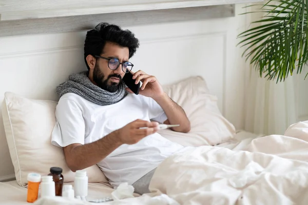 Online Consultation. Sick Indian Man Wrapped In Scarf Holding Thermometer And Calling Doctor On Cellphone While Sitting In Bed At Home, Ill Eastern Man Suffering Cold Or High Fever, Free Space