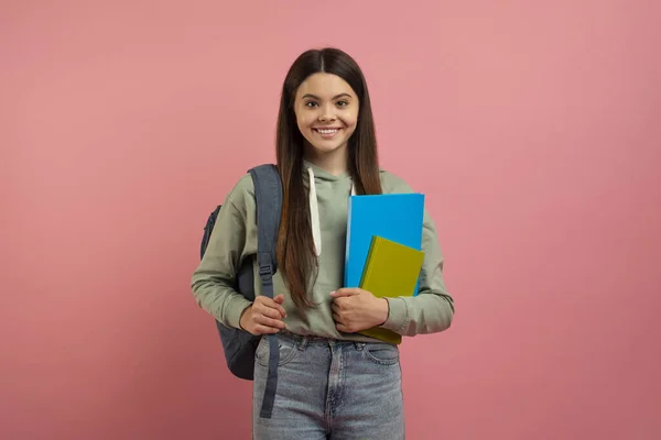 年轻快乐女生的画像 背包和书本装点着粉红的工作室背景 漂亮少女拿着练习本 对着相机微笑 享受现代教育 复制空间 — 图库照片