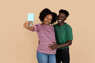 Bin yıllık siyah koca karısına dokunur, hamileliğin tadını çıkarır, akıllı telefondan selfie çeker, bej arka planda izole olur. Gelecekteki aile, yaşam tarzı ve bebek beklentisi, sosyal ağlar için fotoğraf