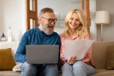 Mutlu bir çift emeklinin bilgisayar kullanması ve ellerinde kağıtlarla evde oturması, üst düzey karı-koca dizüstü bilgisayarlarını okumaları, faturaları kontrol etmeleri, birlikte coşkuyla evrak işleri yapmaları.