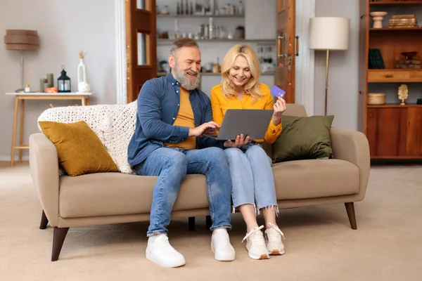 Online Shopping. Happy Mature Couple Using Digital Laptop Computer And Bank Card, Buying And Purchasing Things Sitting On Sofa At Home Interior. Joyful Customers, Sales Offer Concept