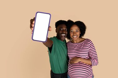 Neşeli siyah koca, karısına sarılıyor, hamileliğinin tadını çıkarıyor, akıllı telefonu boş ekranla gösteriyor, bej arka planda, stüdyoda soyutlanıyor. Aile, bebek, web sitesi, uygulama tavsiyesi