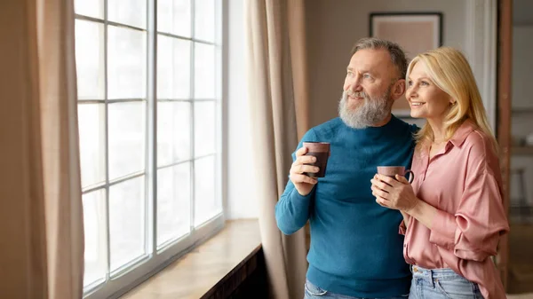Happy Senior Couple Drinking Coffee Hugging Near Window At Home, Looking Aside Enjoying Morning Drink Together At Calmness And Comfort Of Their Own Home Indoor. Panorama With Copy Space