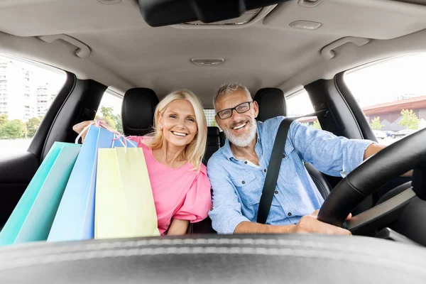 Cheerful beautiful elderly spouses driving home after shopping in mall, smiling. Happy senior man and woman sitting inside car, holding colorful shopping bags purchases. Black friday, season sale