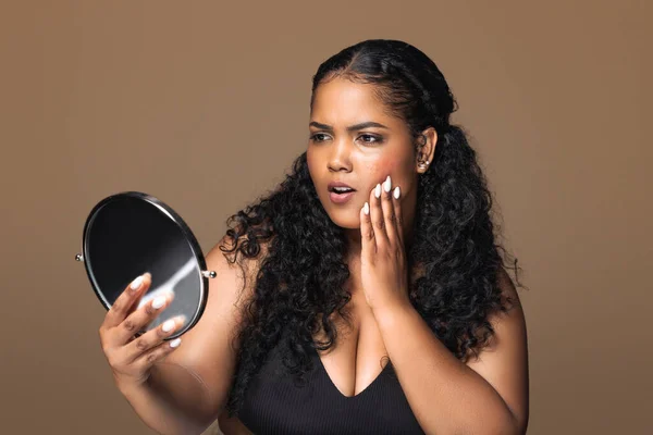 Skin aging concept. Shocked brazilian plus size woman looking at mirror and touching her face, posing over brown studio background, banner