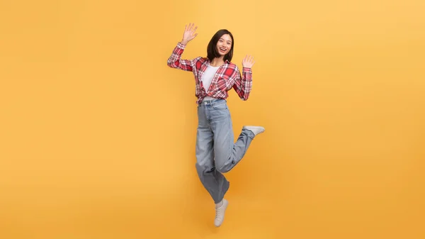 Joyful young asian lady jumping in air and smiling at camera, excited female in casual outfit having fun over yellow studio background, full length, copy space