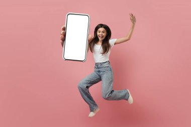 Neşeli genç kız elinde kocaman boş bir akıllı telefonla havaya atlıyor, kaygısız genç kız boş cep telefonu gösteriyor, genç kadın pembe stüdyo arka planında eğleniyor, kolaj, model.