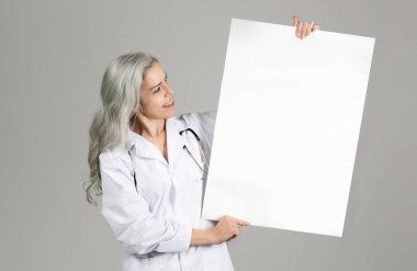 Tıbbi reklam. Orta yaşlı kadın doktor, Gri Stüdyo Arkaplanı Üzerinde Boş Kağıt Posteri, Beyaz Ceketli Poz, Tıp Reklamı Teklifi. Sağlık Reklamları İçin Model