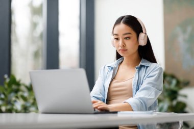 Kulaklık takan Asyalı bayan üniversite öğrencisi dizüstü bilgisayarda online eğitim dersi alıyor, üniversite sınıfında masada oturuyor. Sanal eğitim webinar, uzaklık akademik öğrenim